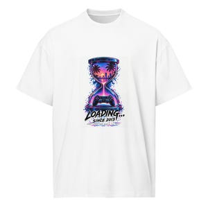 Könnte beinhalten: Ein weißes T-Shirt mit einer Grafik, die eine Sanduhr mit einer Strandlandschaft bei Sonnenuntergang und einem Videospiel-Controller zeigt. Der Text lautet LOADING... SINCE 2013 und SOME GAMES ARE WORTH THE WAIT in leuchtenden Lila-, Blau- und Rosatönen.