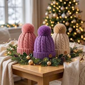 May include: Three cable-knit beanies with pom-poms in dusty pink, vibrant purple, and beige. Each hat features a ribbed cuff and a small circular wooden tag. The beanies are arranged in a row, showcasing their textured knit pattern and soft pom-poms.