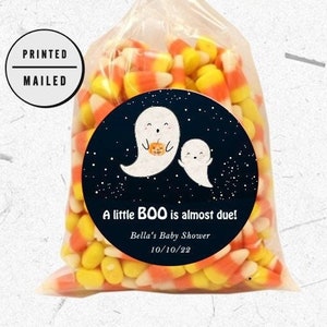 May include: A clear cellophane bag filled with orange and yellow candy corn. A round sticker with two white ghosts and the text "A little BOO is almost due! Bella's Baby Shower 10/10/22" is attached to the bag.