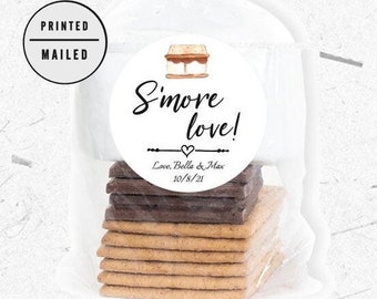 Puede incluir: Una pegatina circular blanca con el texto "S'more love!" y una pequeña ilustración de un s'more. Debajo del texto está "Love, Bella & Max 10/8/21". La pegatina está en una bolsa de plástico transparente llena de galletas graham, cuadrados de chocolate y malvaviscos.