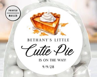 Op de afbeelding: Een witte traktatiezak met een ronde sticker met een aquarel-illustratie van een stuk pompoentaart met slagroom en herfstbladeren. De sticker zegt "Bethany's Little Cutie Pie Is On The Way! 9/9/28".
