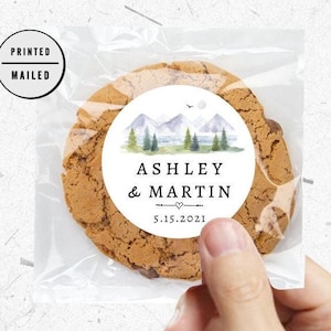 Op de afbeelding: Een ronde sticker met een aquarel-illustratie van bergen, bomen en een meer. De sticker zegt "Ashley & Martin 5.15.2021" en is bevestigd aan een bruine koek.