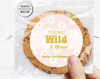May include: A pink and white cookie with a round sticker that says "Young Wild & three" with a daisy pattern. The sticker also says "Bella's 3rd Birthday".