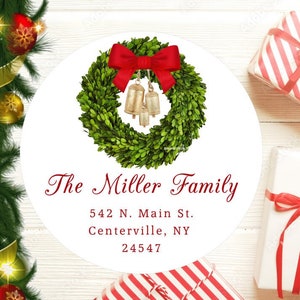 May include: A white round sticker featuring a green boxwood wreath adorned with a red bow and gold bells. The text "The Miller Family" is written in red cursive script. Below the text is the address "542 N. Main St. Centerville, NY 24547" in black font.