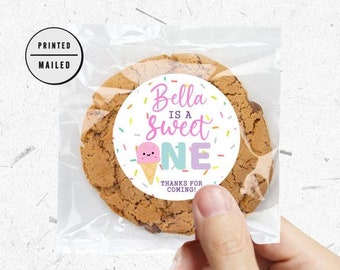 May include: A round sticker with a pink and blue ice cream cone graphic and the text "Bella is a Sweet One" with colorful sprinkles around the edge. The sticker is attached to a brown cookie.