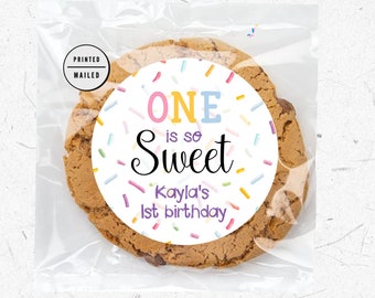May include: A white round sticker with colorful sprinkles and the text "ONE is so Sweet Kayla's 1st birthday" on a brown chocolate chip cookie.