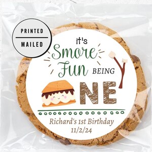Op de afbeelding: Een witte ronde sticker met de tekst "It's Smore Fun Being One" in groene en bruine tekst. De sticker bevat ook een cartoon-marshmallow met een glimlachend gezicht en de tekst "Richard's 1st Birthday 11/2/24".