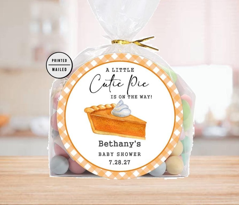 May include: A white and orange favor bag with a round sticker that says "A Little Cutie Pie Is On The Way!" with a picture of a pumpkin pie. The sticker also says "Bethany's Baby Shower 7.28.27".