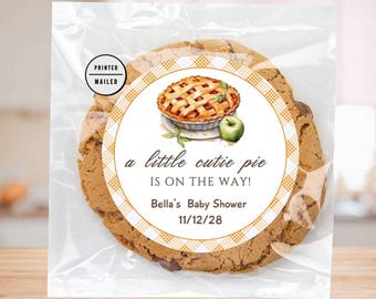 Op de afbeelding: Een ronde koek met een wit en oranje geruit patroon en een sticker met de tekst "a little cutie pie is on the way! Bella's Baby Shower 11/12/28" met een aquarel-illustratie van een appeltaart.