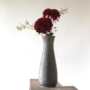 May include: A tall, gray vase with vertical lines holds two large, dark red flowers and delicate green sprigs. The vase sits on a wooden surface against a white background, creating a minimalist floral arrangement.