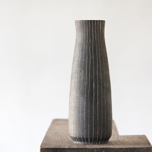 May include: A tall, dark gray vase with vertical, textured lines. The vase has a wide base that tapers to a narrower neck. It is displayed on a wooden surface against a white background. The vase is approximately 12 inches tall.