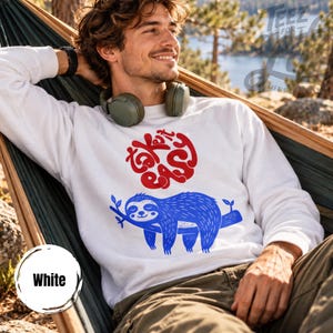 Könnte beinhalten: Ein weißes Langarm-Sweatshirt mit einer roten Grafik, die den Text "take it easy" in einer kreisförmigen Schrift über einer blauen Illustration eines Faultiers zeigt, das an einem Ast hängt. Mit Rundhalsausschnitt und Rippbündchen.