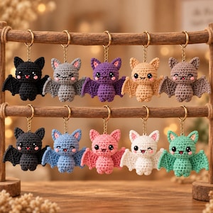 May include: Ten crocheted bat keychains in various colours including black, grey, purple, tan, brown, blue, pink, white, and mint green. Each bat features embroidered eyes, small pink cheeks, and a gold-toned metal ring attached by a short chain.