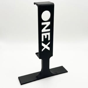 Puede incluir: Un soporte negro vertical en forma de T que presenta una secci&oacute;n vertical rectangular con la palabra ONEX recortada en una fuente sans-serif en negrita. La estructura incluye una base horizontal plana y una peque&ntilde;a secci&oacute;n superior en forma de gancho.