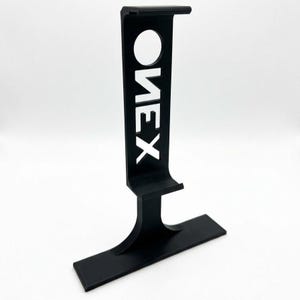 Puede incluir: Un soporte vertical para auriculares en color negro mate con una base rectangular ancha y un brazo de soporte curvo. La secci&oacute;n vertical incluye un recorte circular en la parte superior y la palabra ONEX recortada verticalmente en letra sans-serif.