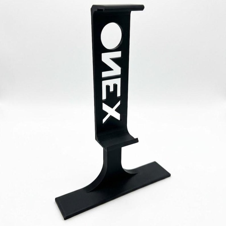 Puede incluir: Un soporte vertical para auriculares en color negro mate con una base rectangular ancha y un brazo de soporte curvo. La secci&oacute;n vertical incluye un recorte circular en la parte superior y la palabra ONEX recortada verticalmente en letra sans-serif.