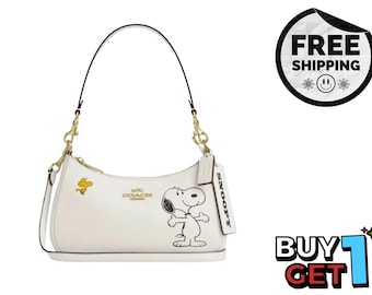 May include: A white leather shoulder bag featuring a graphic of Snoopy and Woodstock. The bag has gold-tone hardware, a zipper closure, and a detachable leather tag embossed with the name Snoopy. The Coach logo is printed in gold on the front.