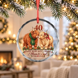 Könnte beinhalten: Ein runder, klarer Glasanhänger mit zwei Highland-Rindern, eines mit grünem Schal und das andere mit roter Schleife. Der Text lautet 40, Happy Ruby Anniversary, Mum & Dad und 1986-2026. Er hängt an einem roten Band vor unscharfem Hintergrund.