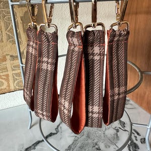 May include: Five fabric wristlet keychains featuring a brown and tan plaid pattern on the exterior and a solid burnt orange lining. Each keychain is attached to a polished gold-tone metal lobster clasp, designed for securing keys or small accessories.
