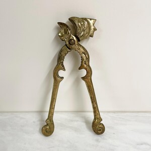 May include: A brass nutcracker featuring a sculpted eagle head at the top hinge. The tool has two long, textured handles that curve into spiral ends at the base. The metal surface shows a weathered, golden-toned patina with intricate detailing.