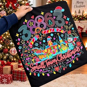 May include: A black fabric item featuring a neon-style graphic with the number 38863, a gingerbread person, a snowman, and a small town scene. The text reads Small Town Christmas PONTOTOC, MS, surrounded by colourful holiday lights and stars.