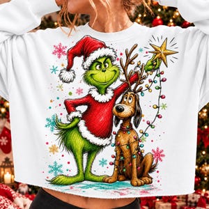 Può includere: Una felpa bianca a maniche lunghe con una stampa grafica di un personaggio verde in costume da Babbo Natale rosso e un cane marrone con le corna. Il personaggio tiene in mano una stella dorata e una catena di luci natalizie colorate.