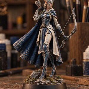 May include: A detailed figurine of an elf archer standing on a rocky base. The figure wears blue and cream armour with a matching cape, a pointed helmet, and carries a bow in one hand and a quiver of arrows on the back, featuring intricate gold trim.