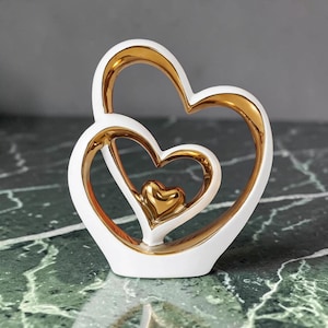 Op de afbeelding: Een decoratief beeldje met drie in elkaar grijpende hartvormen. De buitenste en middelste harten hebben een gladde witte buitenkant met een reflecterende gouden binnenkant, terwijl het kleinste hart volledig goudkleurig is.