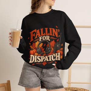 May include: A black long-sleeve sweatshirt featuring a graphic print with the text "FALLIN FOR DISPATCH" in orange and cream. The design includes a headset, a 911 radio, and orange autumn leaves and pumpkins on a dark background.