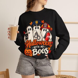May include: A black long-sleeve sweatshirt featuring a graphic of three ghosts dressed as a police officer, a firefighter, and a dispatcher. The text reads "Halloween WITH MY Boos" in orange and white, surrounded by autumn leaves and a pumpkin.