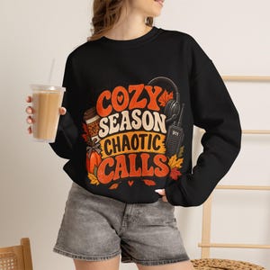 May include: A black long-sleeve sweatshirt featuring a graphic print with the text "COZY SEASON CHAOTIC CALLS" in orange and cream. The design includes illustrations of a coffee cup, a pumpkin, headphones, and a two-way radio with "911" on it.