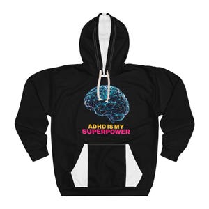 Può includere: Una felpa nera con cappuccio che presenta una grafica di un cervello umano blu e luminoso con piccoli punti bianchi. Sotto il cervello, il testo recita ADHD IS MY in giallo e SUPERPOWER in rosa. La felpa include una tasca a marsupio e lacci bianchi.