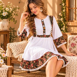 May include: A white dress featuring black floral panels on the bodice, sleeves, and hem. The garment is accented with white pom-pom trim along the edges of the floral sections and includes a round neckline with three central white buttons.