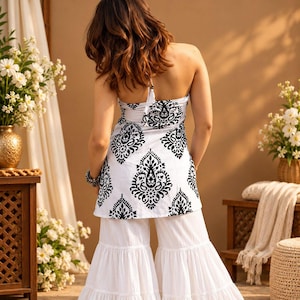 May include: A white halter-neck top featuring a black paisley-inspired print, paired with white wide-leg trousers that have a tiered, ruffled hem. The top has a tie closure at the back of the neck and a fitted bodice that flares slightly at the waist.