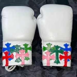 Pode incluir: Um par de luvas de boxe brancas com uma série de remendos costurados em forma de cruz nas cores azul, verde, vermelho e rosa ao redor dos punhos. As luvas têm uma superfície lisa e são projetadas com um fecho de cadarço na base.