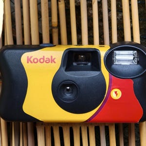 Op de afbeelding: Een Kodak wegwerpcamera met een zwarte, gele en rode behuizing. De voorzijde toont het Kodak-logo in rode tekst, een centrale lens, een rechthoekige zoeker, een flitser en een kleine gele ronde knop met een rood bliksemschichtpictogram.