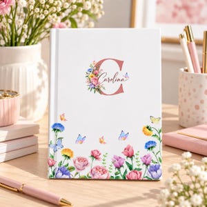 Puede incluir: Un diario de tapa dura blanca con una letra C grande de color rosa y el nombre Carolina escrito en cursiva. La portada está decorada con coloridas ilustraciones en acuarela de varias flores y mariposas a lo largo del borde inferior.