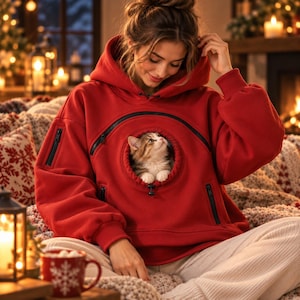 May include: A bright red hooded sweatshirt featuring a large, circular front pouch with a black zippered opening. A small, fluffy calico kitten is visible inside the pouch, looking upward. The sweatshirt includes long sleeves with ribbed cuffs and additional black zippered pockets on the sides.