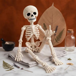 May include: A crocheted off-white skeleton figure with black yarn detailing around the eye sockets and teeth. The figure is posed in a seated position with one hand raised, featuring a visible rib cage, pelvic structure, and articulated limbs.