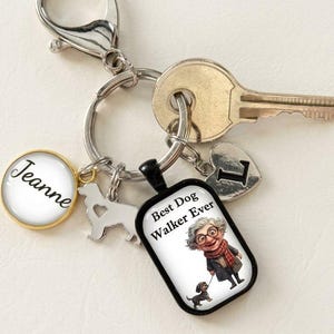 May include: A silver-toned keychain featuring a metal ring with a lobster clasp, a round charm with the name Jeanne, a dog-shaped charm, a heart charm with the letter L, and a rectangular charm displaying an illustration of a person walking a dog.