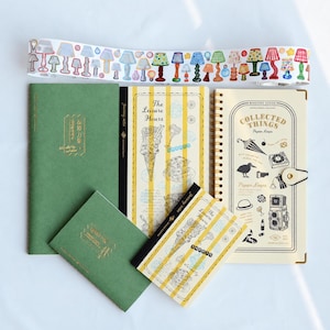 May include: A collection of stationery including two green notebooks with gold foil text, two yellow striped notebooks titled The Leisure Hours, a spiral-bound cream notebook titled Collected Things Paper Keeps, and a roll of decorative lamp washi tape.
