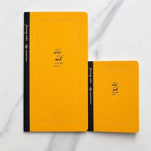 May include: Two mustard yellow planners with black spines, featuring the text Journey notes, 2027, and Monthly Plan on the front covers. The covers have a debossed rectangular border and a small diamond-shaped logo at the bottom center.