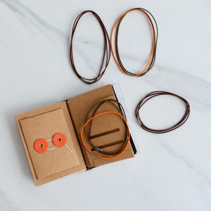 May include: A brown leather notebook with a flap closure secured by two orange circular fasteners, containing internal card slots. Three thin cord necklaces in brown, orange, and olive green are arranged around the open notebook.