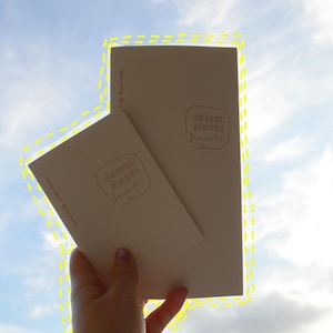May include: Two off-white paper notebooks of different sizes, each featuring the text "TRAVEL NOTES" and "freewriter" enclosed in a hand-drawn rectangular border. The notebooks are held against a bright, cloudy sky with a yellow dashed outline.