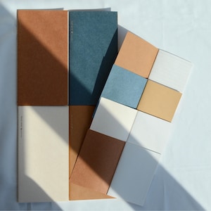 May include: A collection of rectangular notebooks and paper pads in various shades of brown, blue, and cream. The items feature solid-colored covers and interior pages with lined, grid, and blank layouts. Some covers display small printed text.