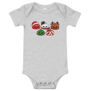 Peut inclure: Un body pour bébé gris chiné à manches courtes avec une impression graphique de cinq personnages ronds, mignons et souriants, portant des accessoires sur le thème des fêtes, dont un bonnet de Père Noël, un chapeau de bonhomme de neige et des bois.