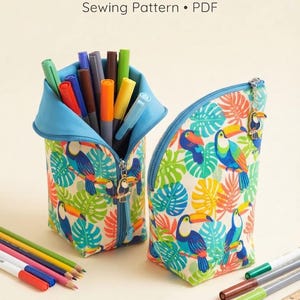 May include: A stand-up pencil case sewing pattern PDF featuring a toucan and tropical leaf print. The case is shown open with markers inside and closed with a toucan zipper pull. The fabric has a cream background with blue, green, and orange motifs.