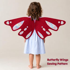 May include: Quilted butterfly wings in deep red fabric featuring floral patterned cutouts with light blue, pink, and white accents. The wings are worn over a light blue pinstriped dress. Text in the bottom right corner reads Butterfly Wings Sewing Pattern.