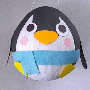 May include: A white penguin pinata with black wings, a blue scarf, and yellow feet. The penguin has a black and white face with orange beak and pink cheeks.
