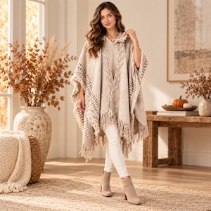 May include: A beige hooded poncho featuring a textured chevron pattern along the front and sides, finished with long fringe trim along the hem and edges. The garment is worn over white slim-fit trousers and beige ankle boots with a block heel.